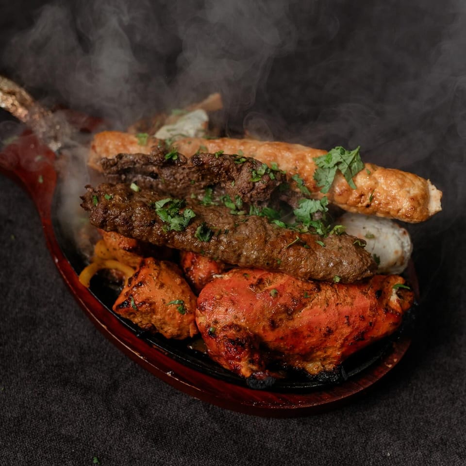 Steaming Indian mixed grill platter with tandoori chicken, seekh kebabs, and other meats on a wooden dish.