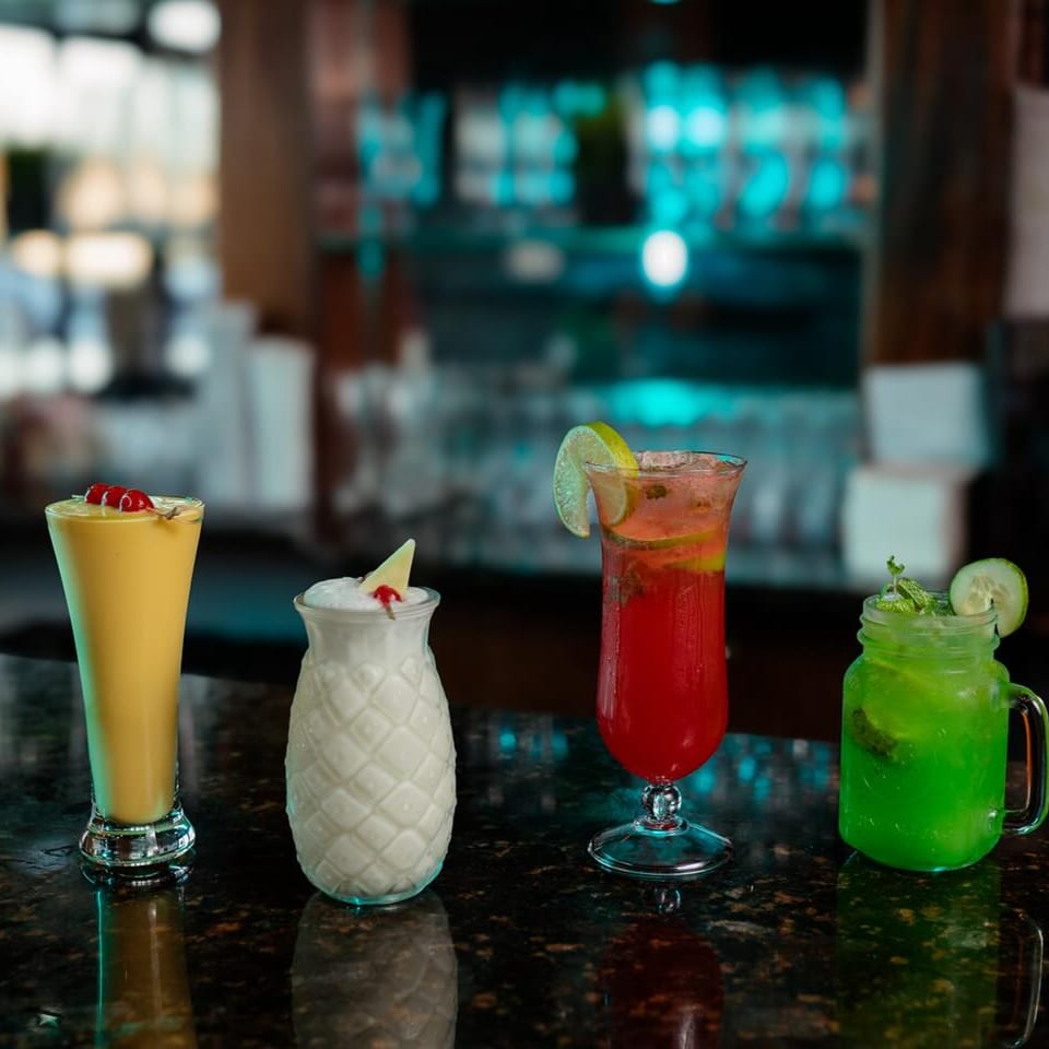 Four colorful drinks, including a yellow lassi, on a dark bar counter at Woondal Restaurant.
