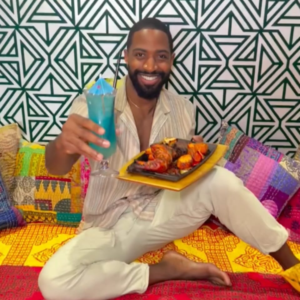 A man smiling, holding a blue drink and a platter of Indian grilled meats and vegetables.